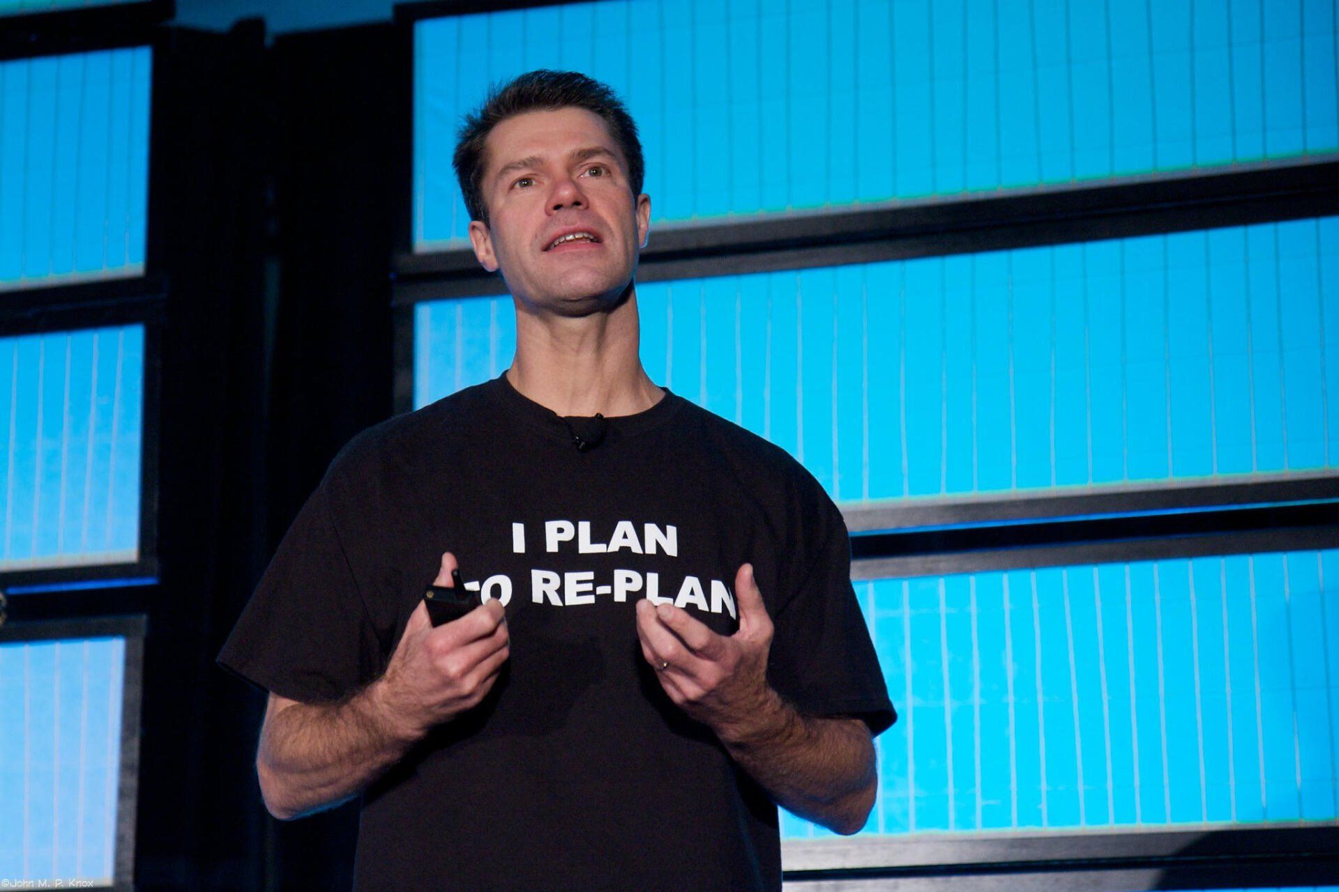 A shot of Luke Hohmann, a white man with short dark hair, giving a talk on stage. He is wearing a black t-shirt that reads 