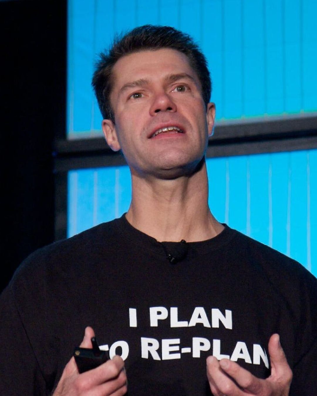 A close up photo of Luke Holmann while he speaks on a stage. Luke is a white man with brown hair and is wearing a black t-shirt that reads 