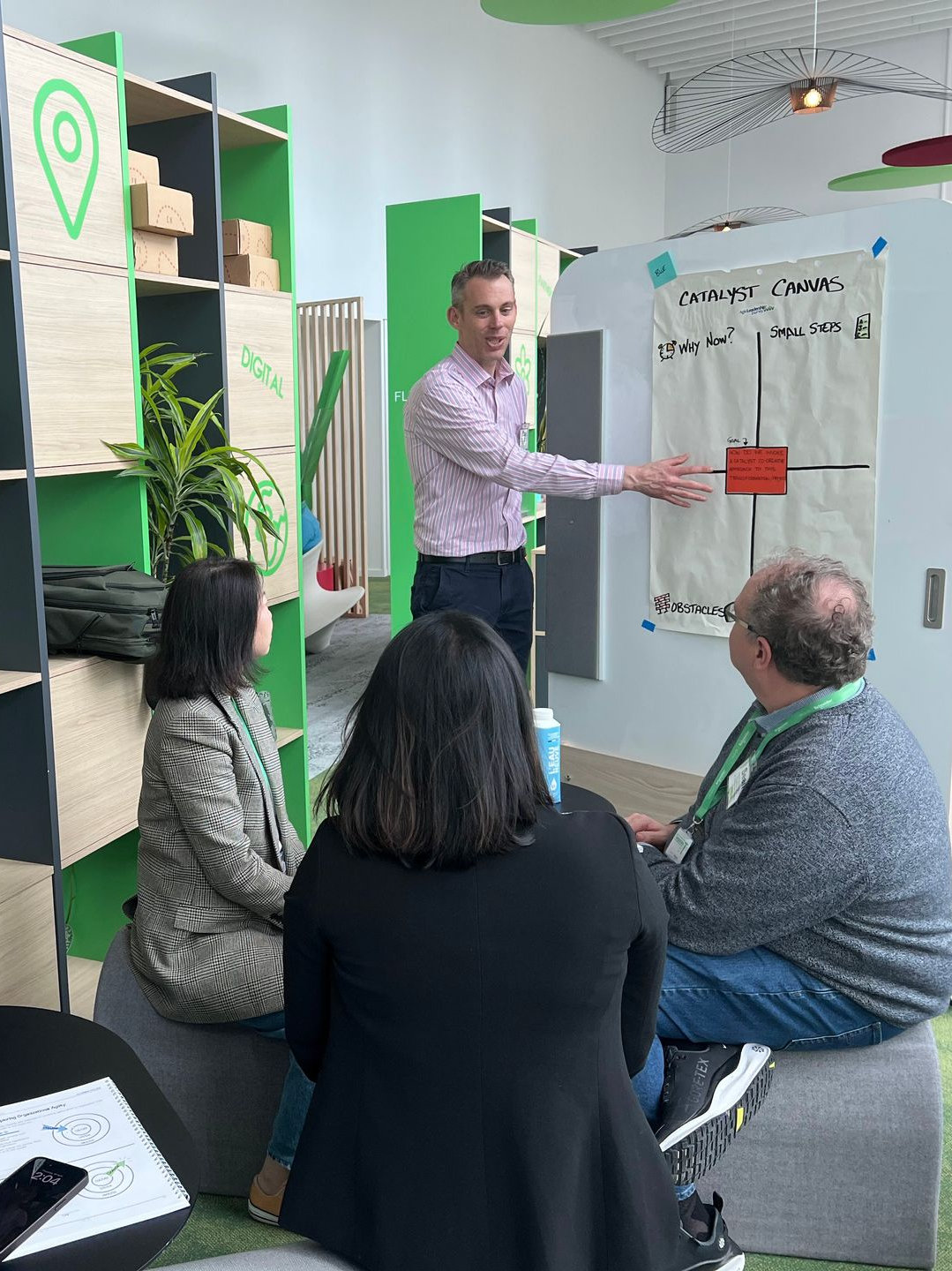 A person in a pink shirt presents to seated people using a quadrant chart on a wall in a bright office space.