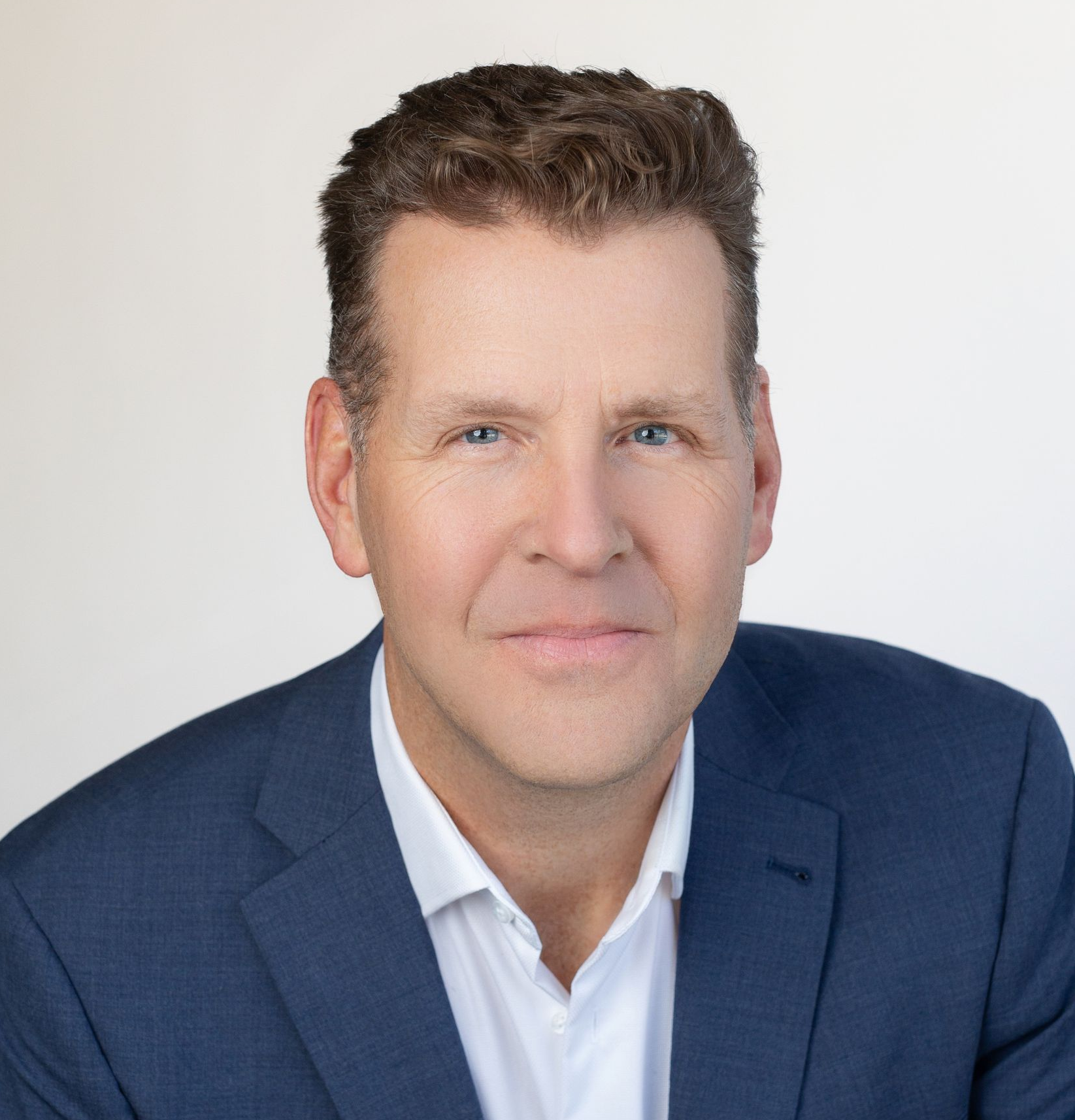 Man in a blue suit and white shirt, smiling, headshot against a white backdrop.