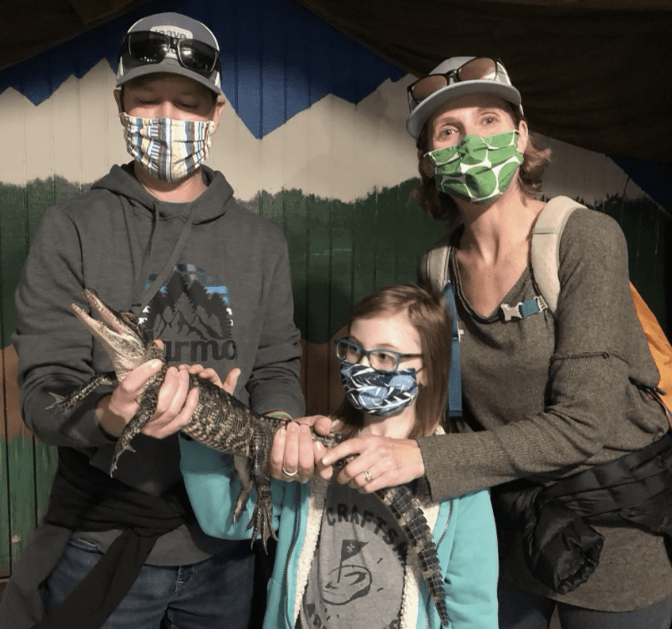 Two adults and a small girl, all wearing face masks, hold an alligator.