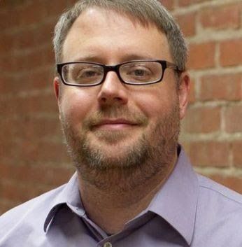 A man wearing glasses and a purple shirt is standing in front of a brick wall.