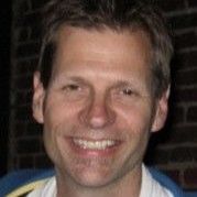 Man with fair skin smiles at the camera, wearing a light-colored shirt in front of a brick wall.