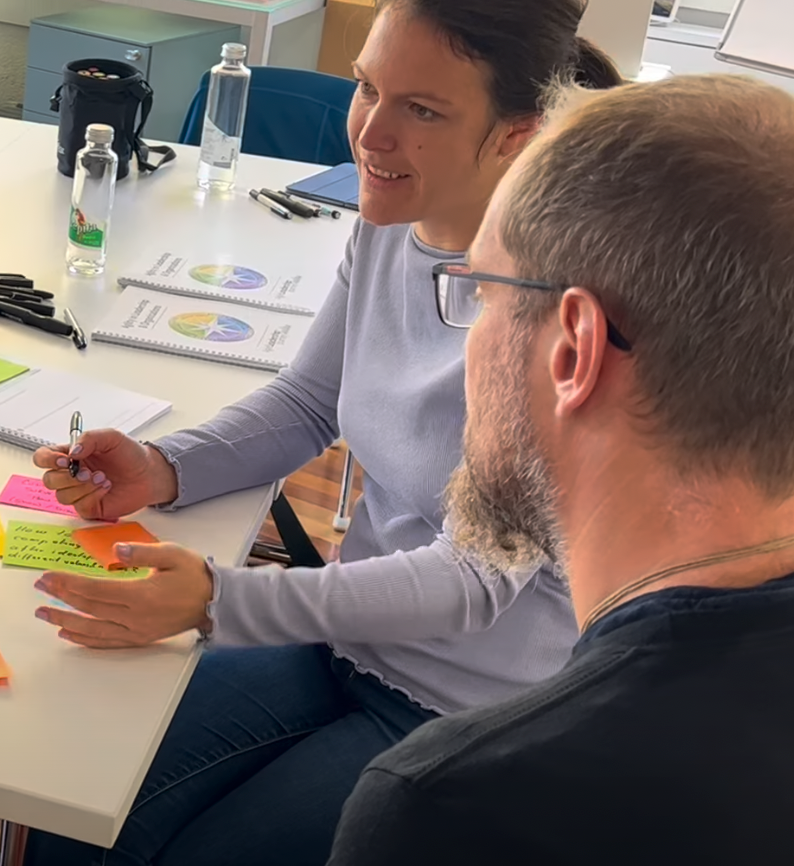 Woman gesturing and speaking to a man at a table with papers and sticky notes; natural light.