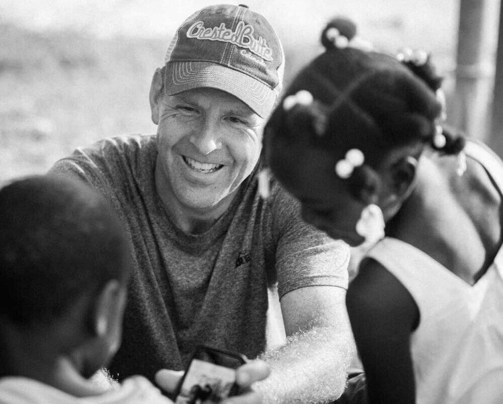 A man, Pete Behrens, wearing a hat is smiling while talking to two children.