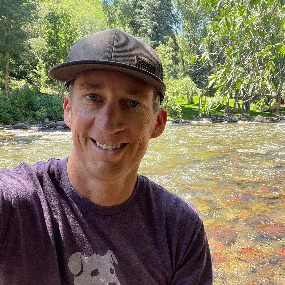 A selfie of Jon Christensen, a light skinned male. Jon is wearing a baseball hat and a t-shirt with a dog on it. A creek is visible in the background.