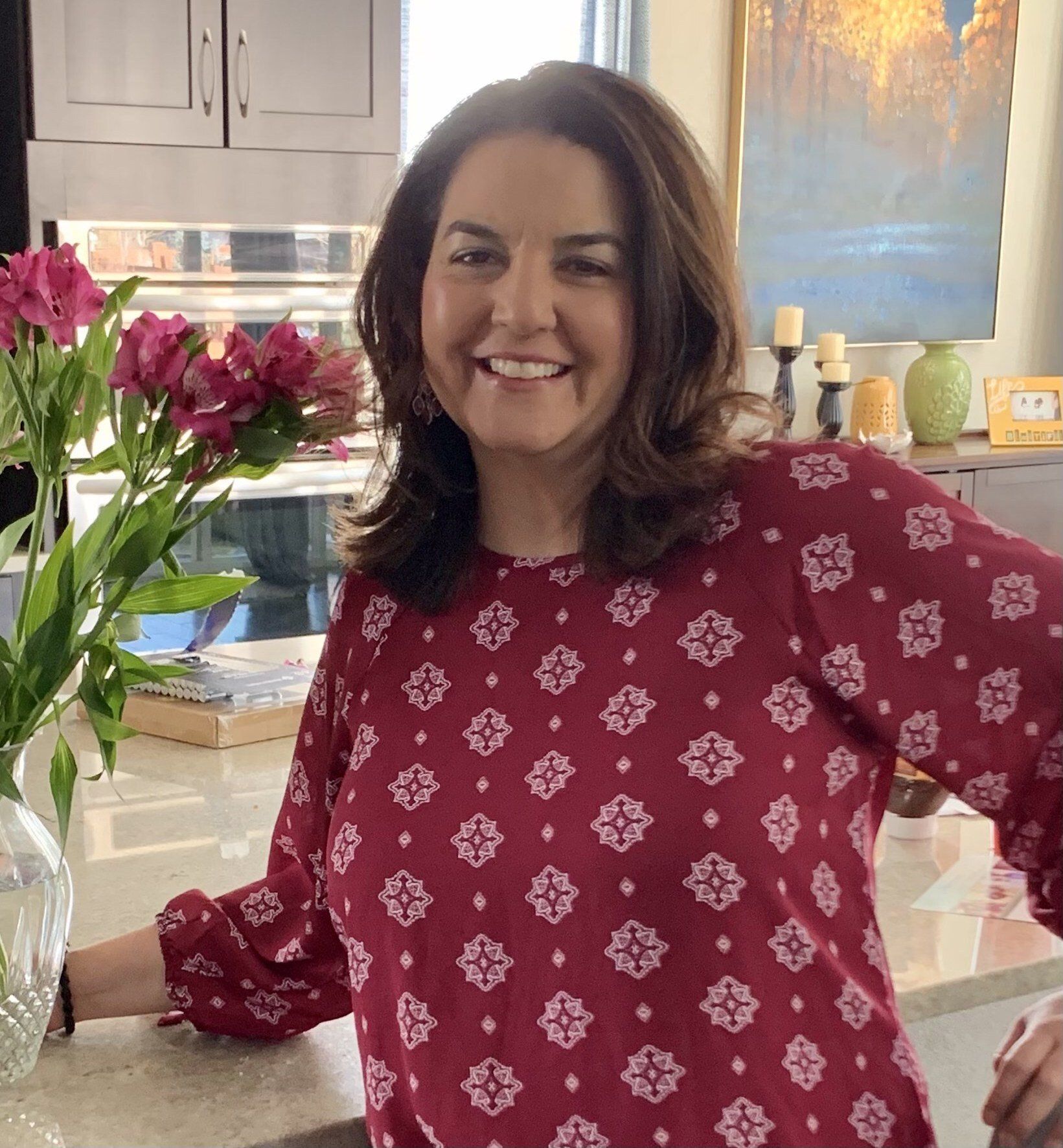 A close up photo of Melissa Uribes, a lighter skinned woman with shoulder length brown hair. Melissa is wearing a pinky-red top with a white pattern printed on it. a vase of flowers can be seen to her right.