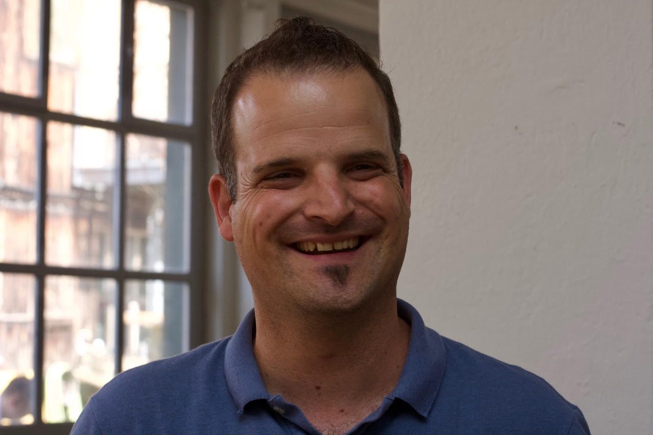 A man in a blue shirt is smiling in front of a window.