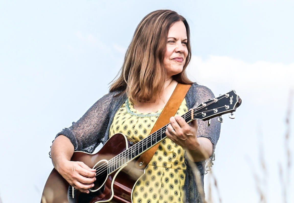A woman named Joy Zimmerman in a yellow dress is holding a guitar in a field.