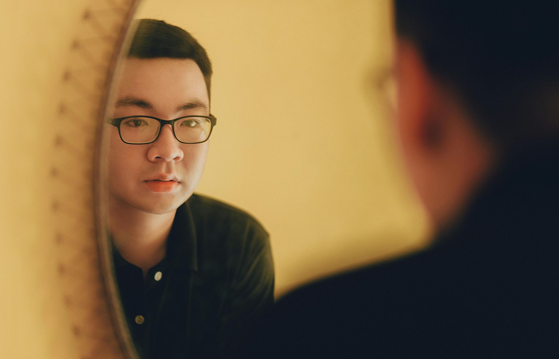 Man with glasses looks at his reflection in a round mirror; warm light.