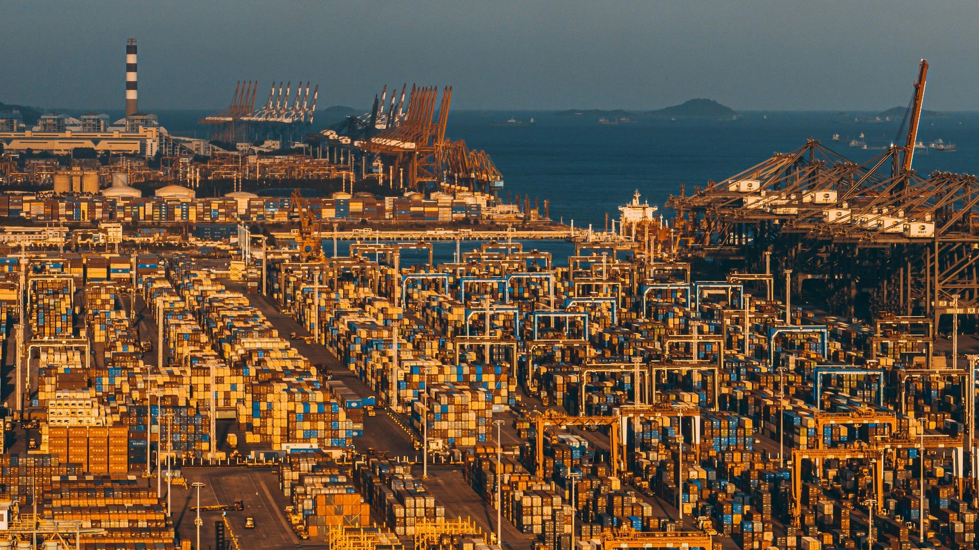 A vast industrial shipping port at sunset, packed with cargo containers, gantry cranes, and machinery near the ocean.