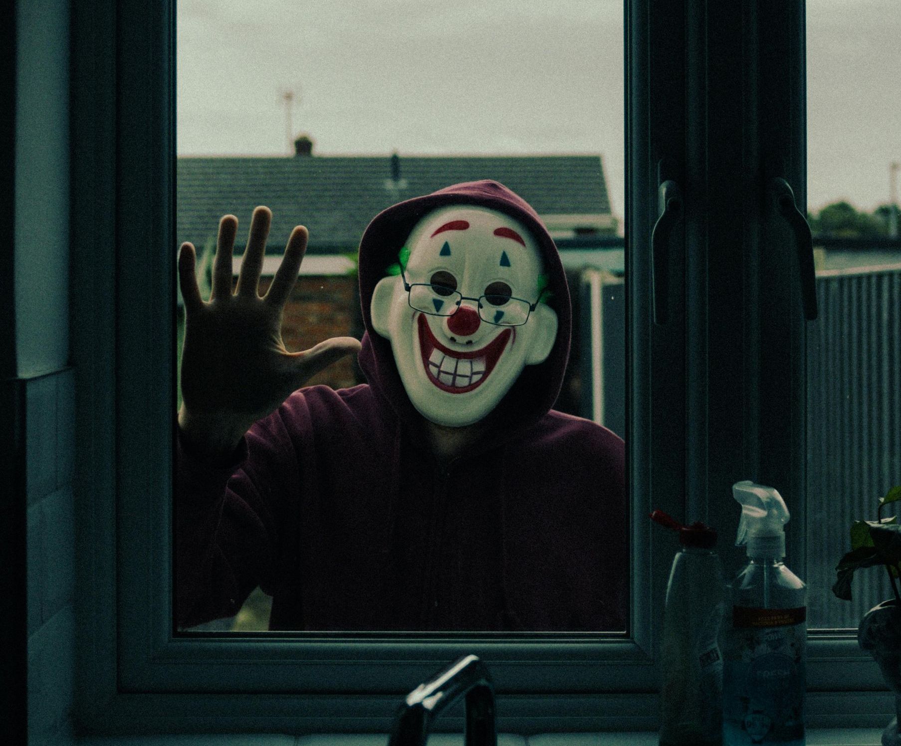 A person wearing a clown mask is looking out a window