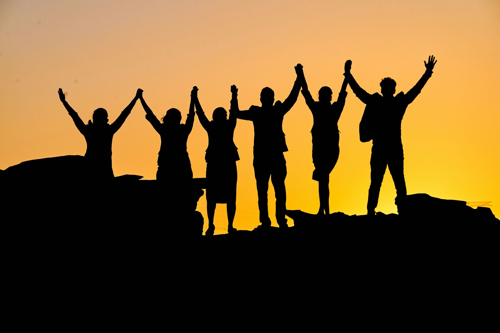 A group of people standing together on a ridge at sunset with arms raised, symbolizing shared achiev