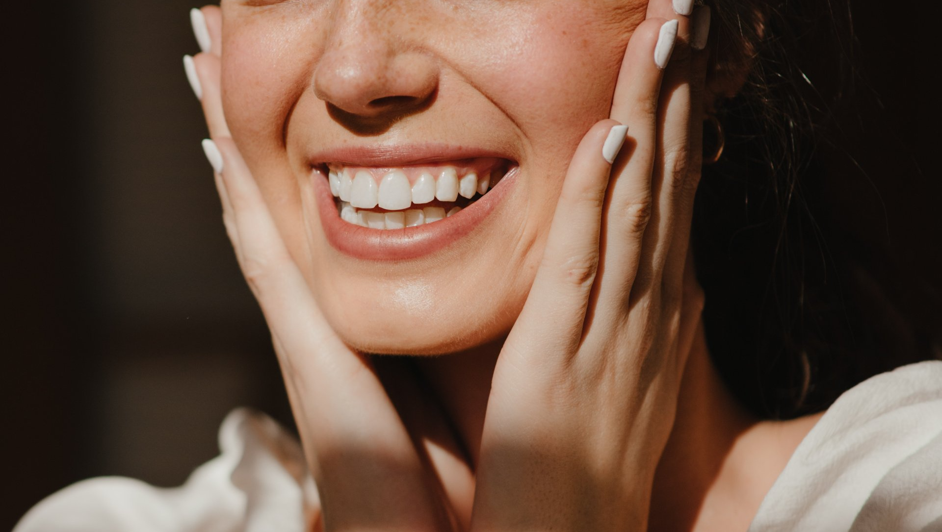 A close up of a woman smiling, full teeth, with nose and chin visible. Her hands are placed on either cheek