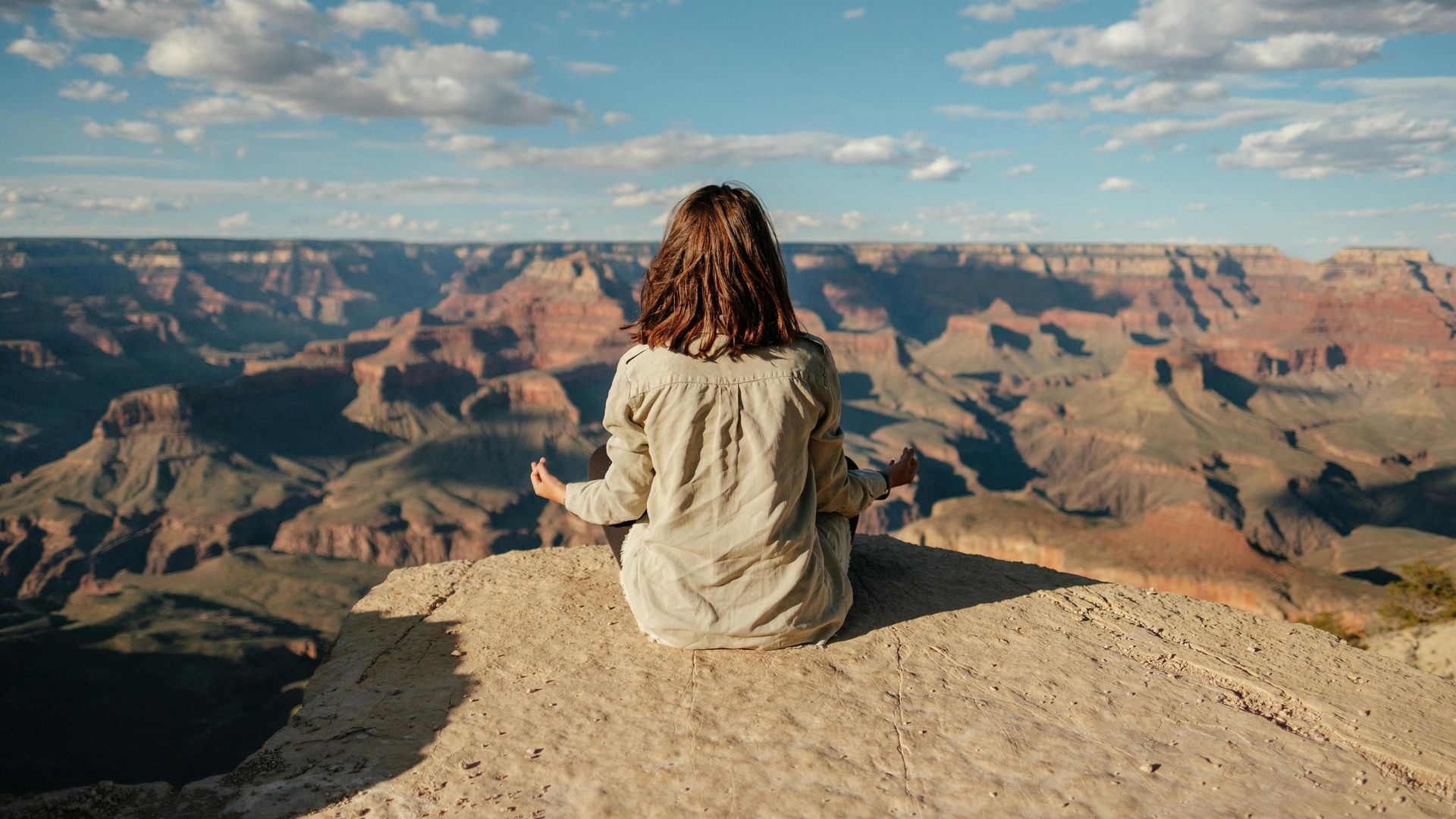 A person meditates in a cross-legged position on the edge of a rocky cliff overlooking a vast, expansive canyon landscape.