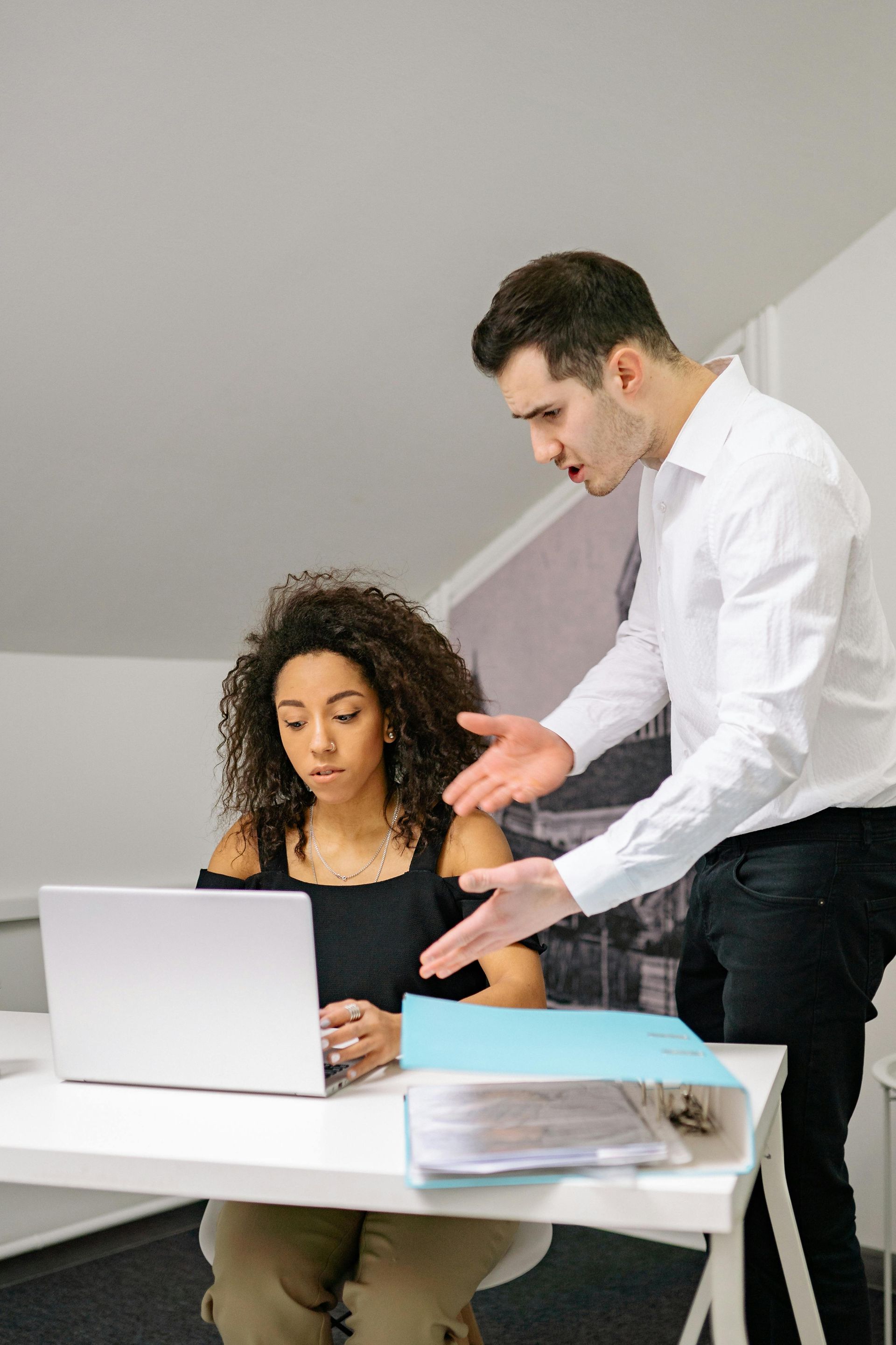 A man is standing next to a woman sitting at a desk using a laptop computer.