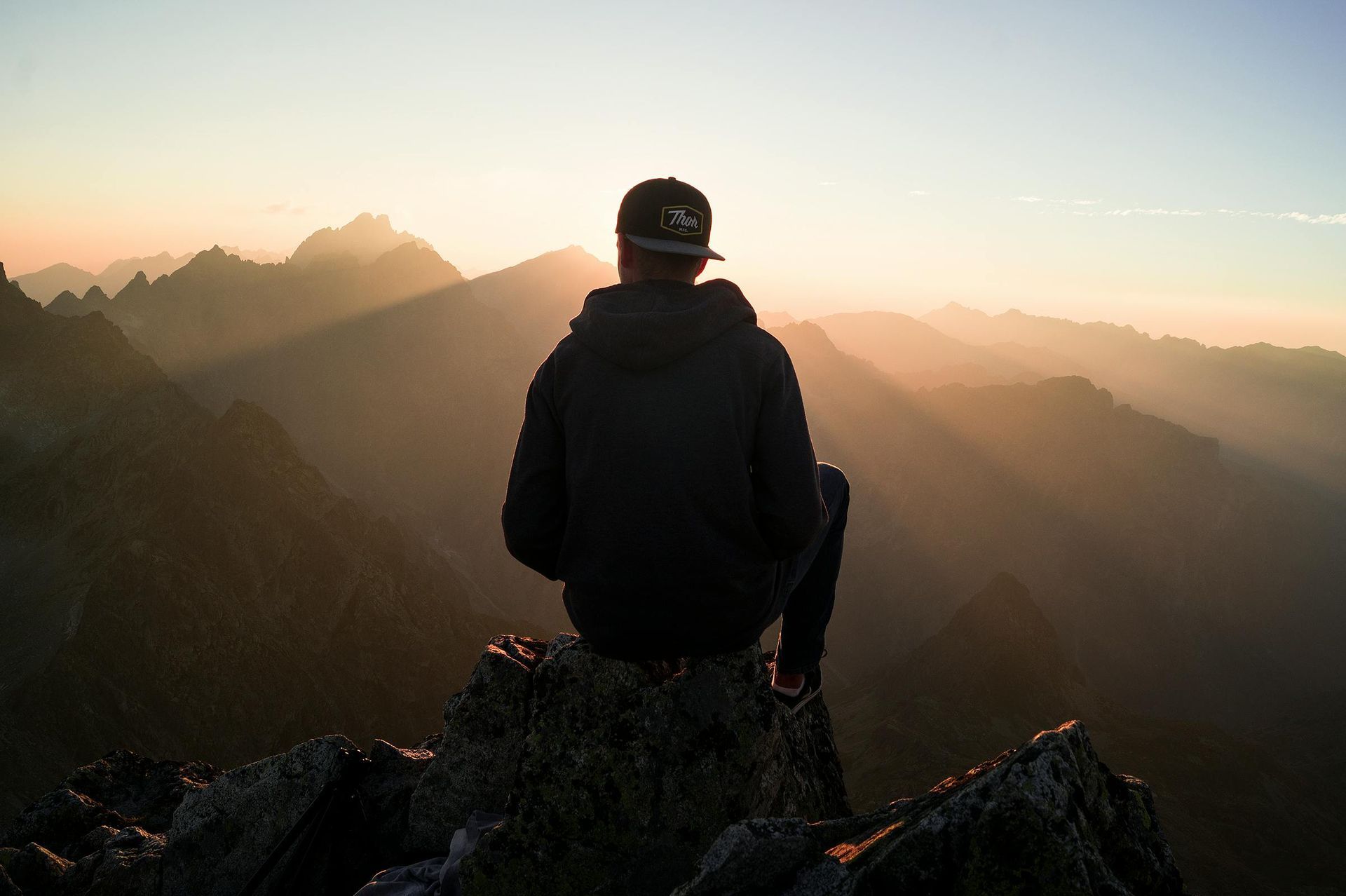 Person in hoodie sits atop a mountain peak, overlooking sunlit mountain range.