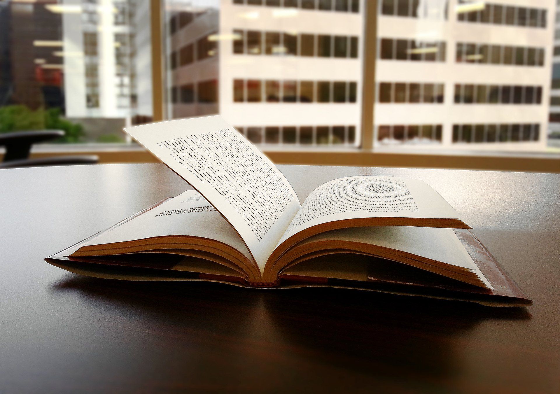 An open book on a table. A high-rise building is visible through the window in the background.