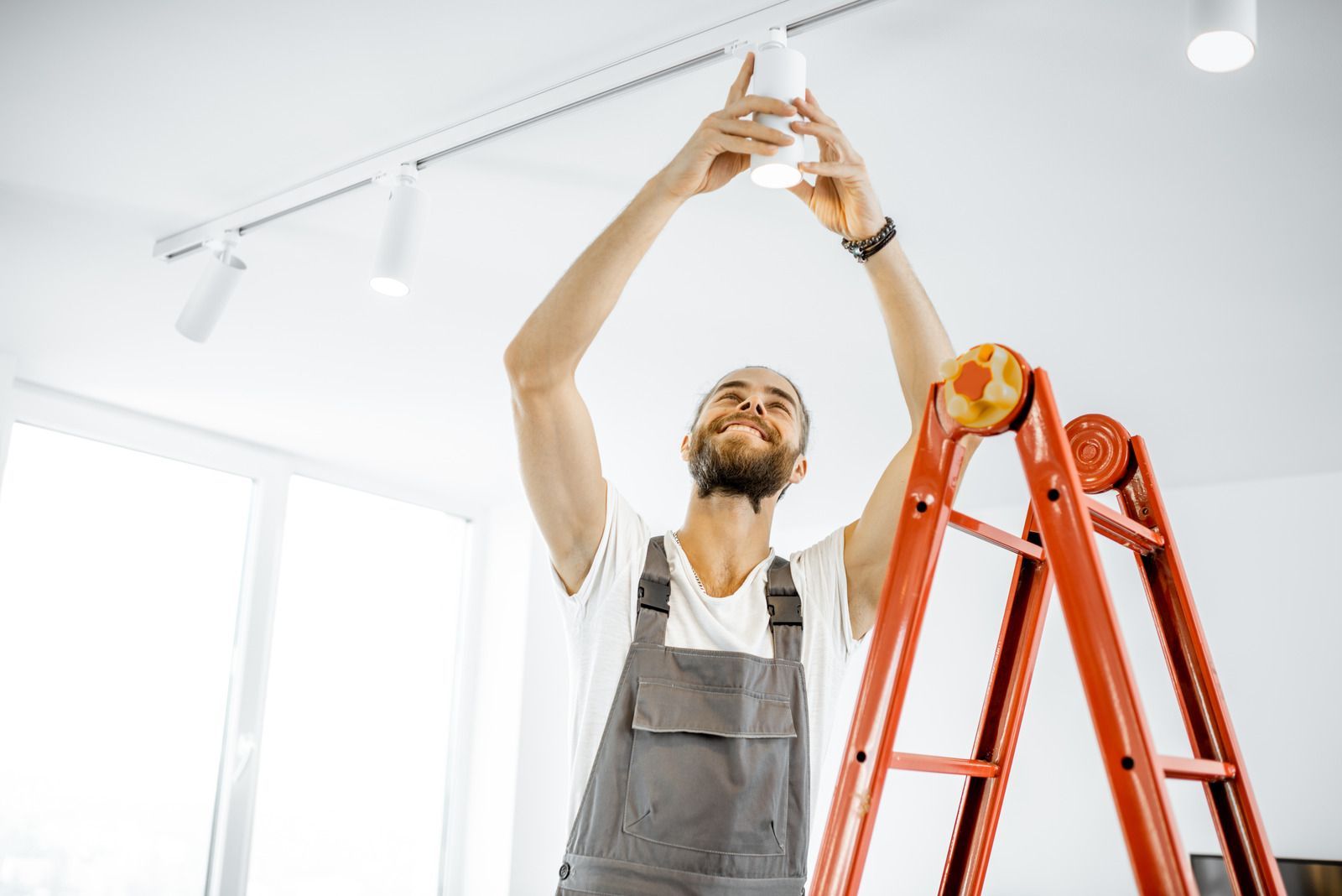 Man in overalls replacing a light bulb on a ladder indoors.