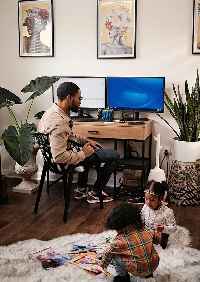 Man working at desk with two monitors while two young children play on a rug.