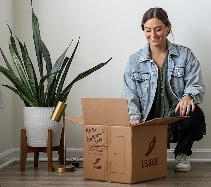Woman in denim jacket smiles as she opens a moving box labeled