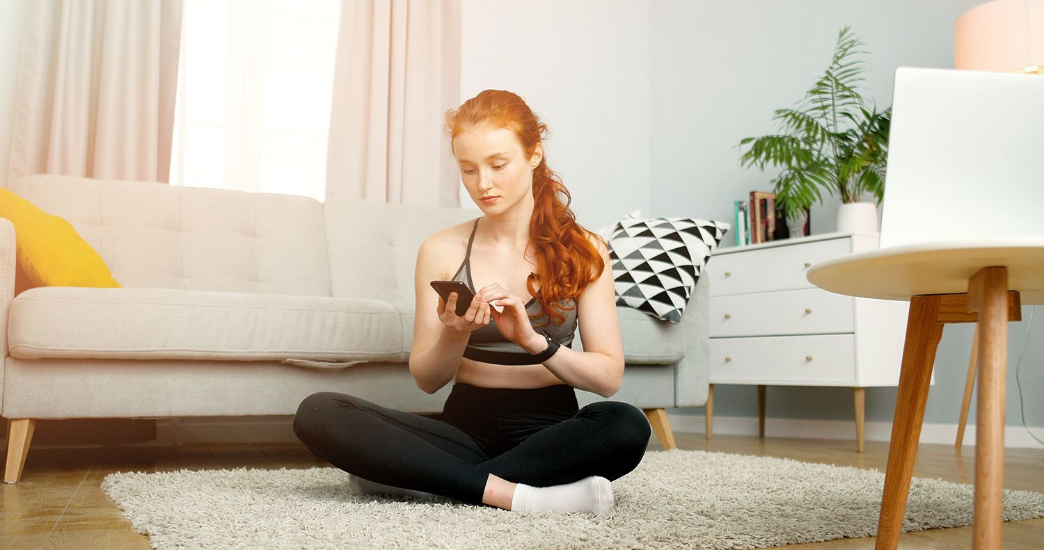 Woman with red hair in workout clothes using phone, sitting cross-legged on rug, near a laptop and couch.