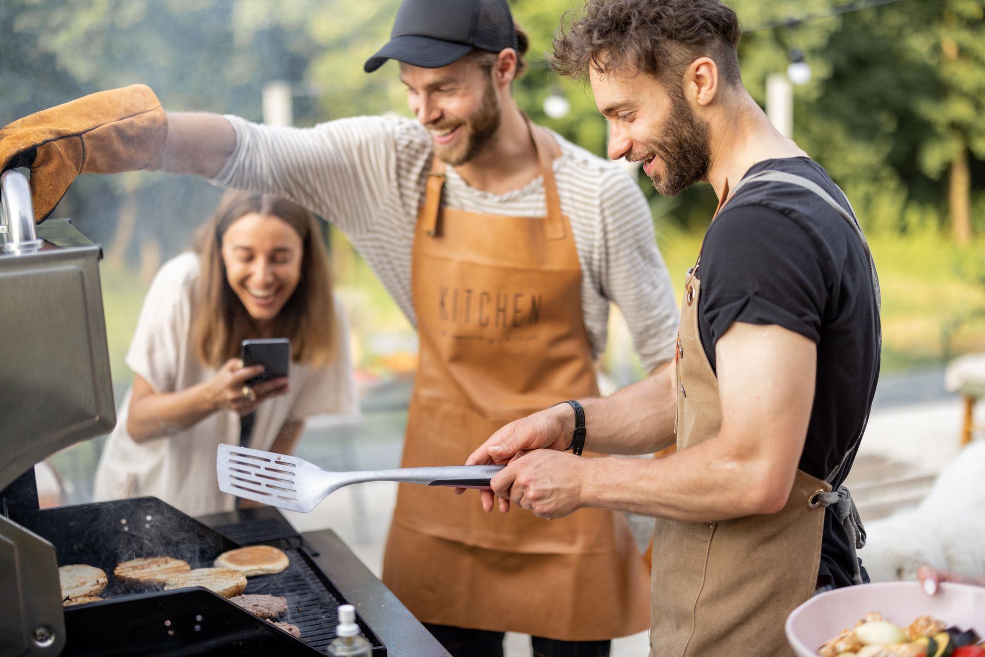Three people at a grill: Two men in aprons cooking, woman taking a photo; outdoors, sunny.