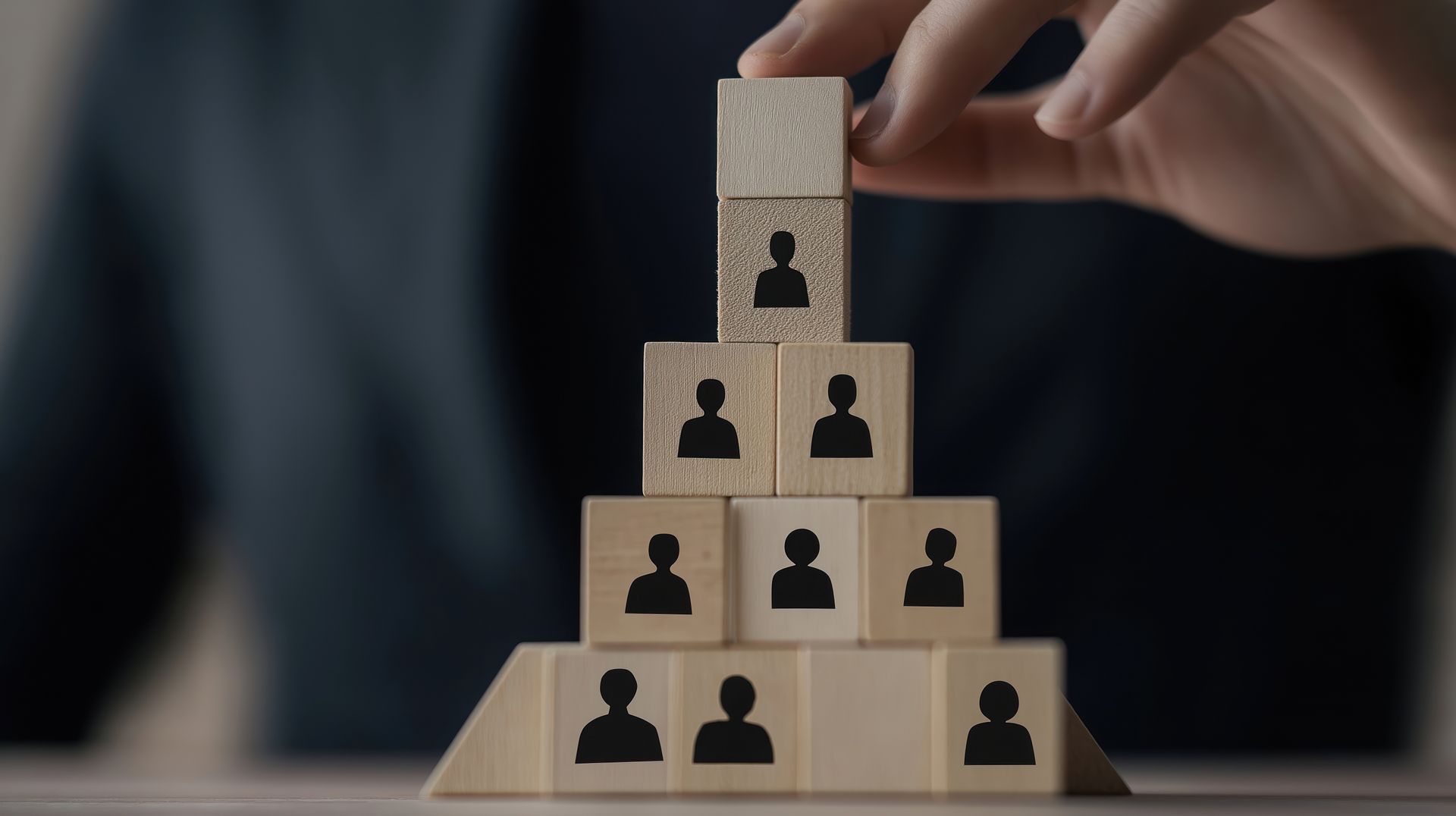 A hand adds a wooden block with a person icon to the top of a pyramid structure, representing organizational hierarchy.