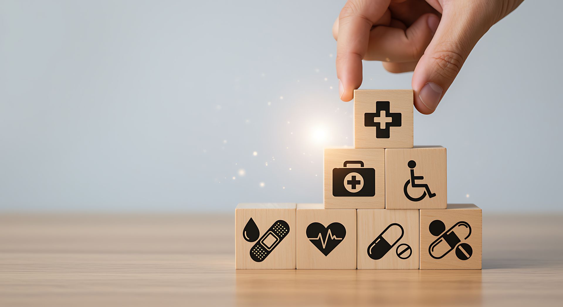 A hand places a wooden block with a medical cross on top of a pyramid stack of health-related icon blocks.