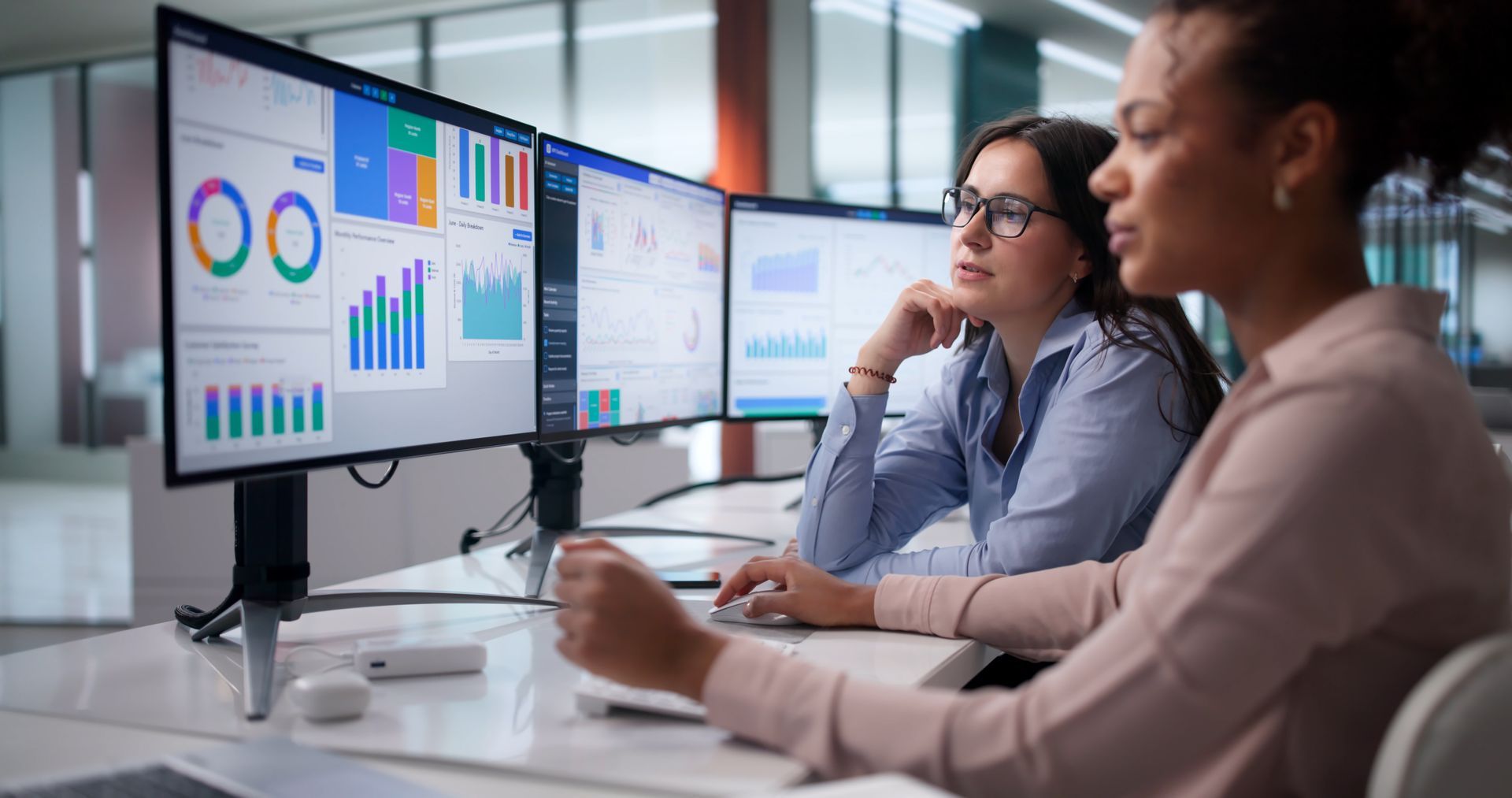 Two professionals collaborate at a desk with multiple monitors displaying data charts and analytics in a bright office.