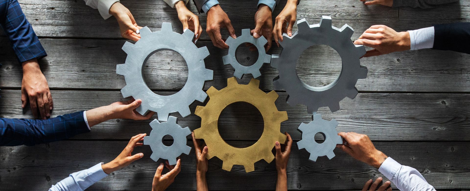 Multiple pairs of hands around a rustic wooden table arrange several gray and gold interlocking gears.