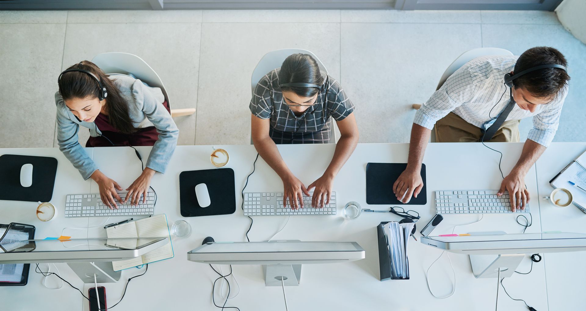 Overhead view of three people sitting at a white desk, each working on their own computer with keyboards and mice.
