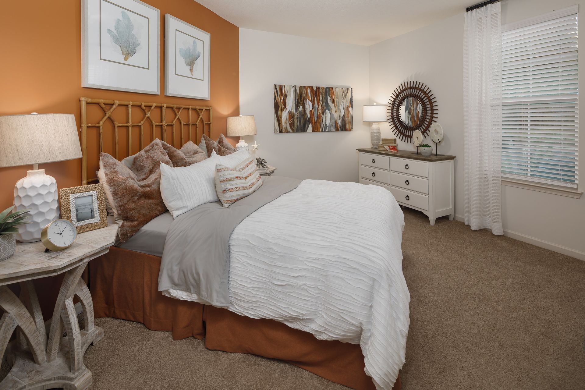 Bedroom with orange accent wall, white bedding, and neutral carpet. Includes a dresser, bedside table, and artwork.