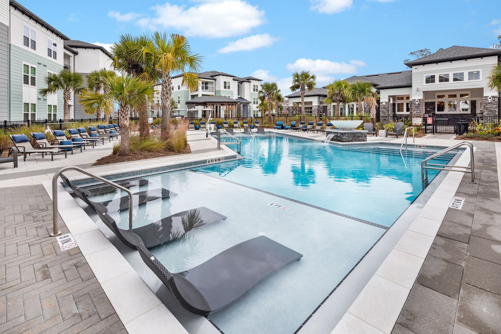 A pool with in-water lounge chairs in front of apartment buildings, under a bright blue sky.