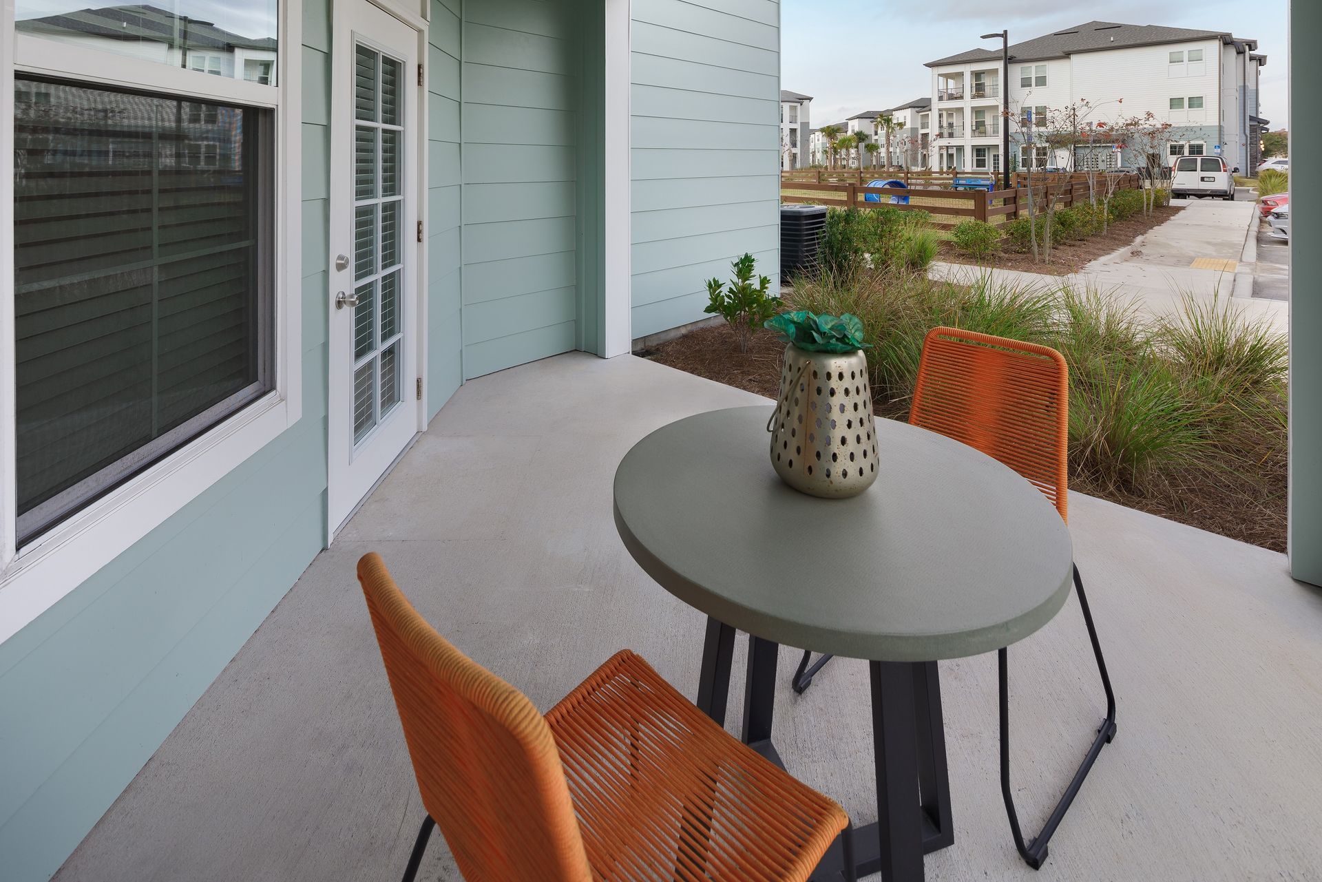 Patio with round table, orange chairs, and decorative jar. Buildings and greenery in the background.