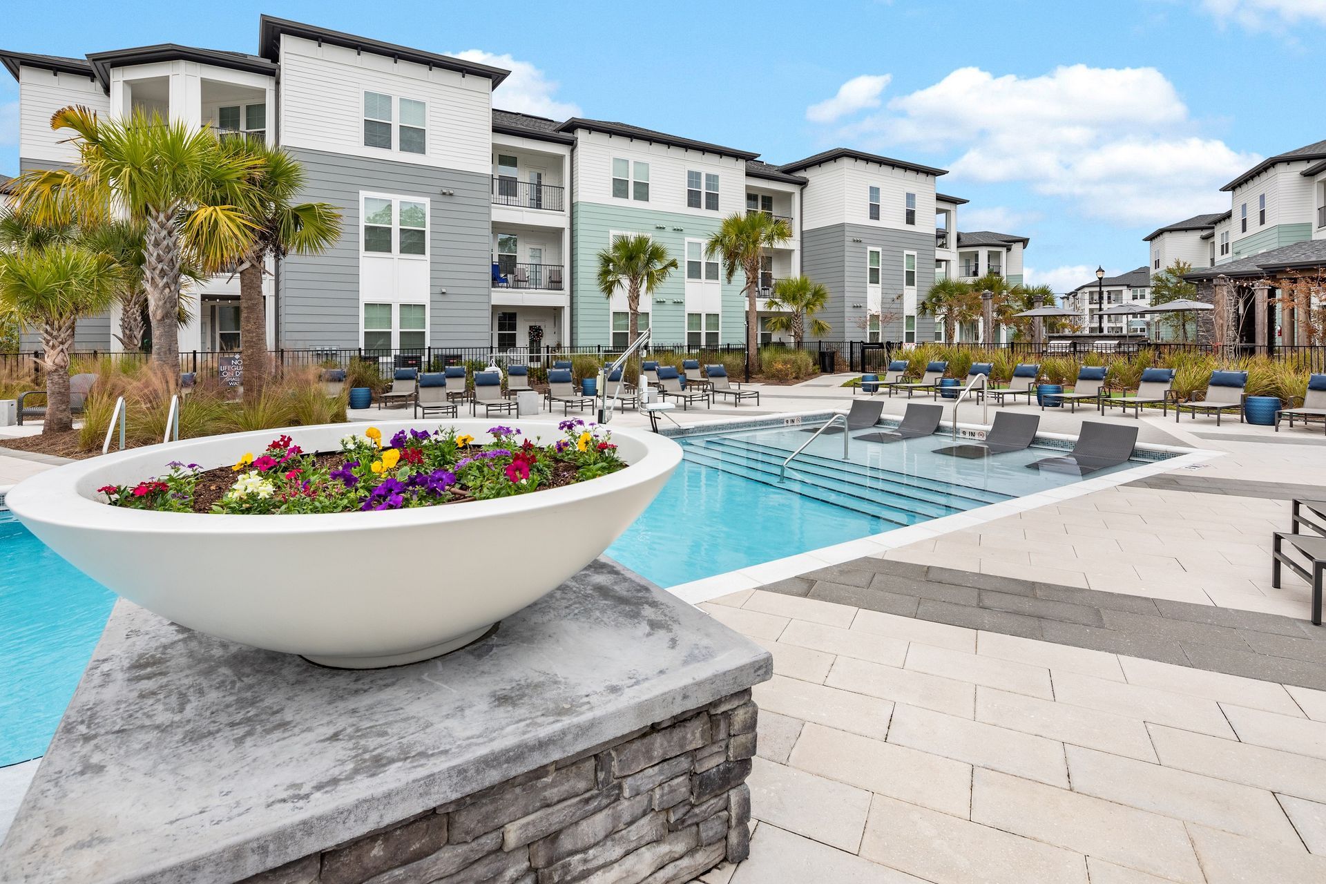 Pool with lounge chairs, flower planter, and apartment buildings under blue sky.