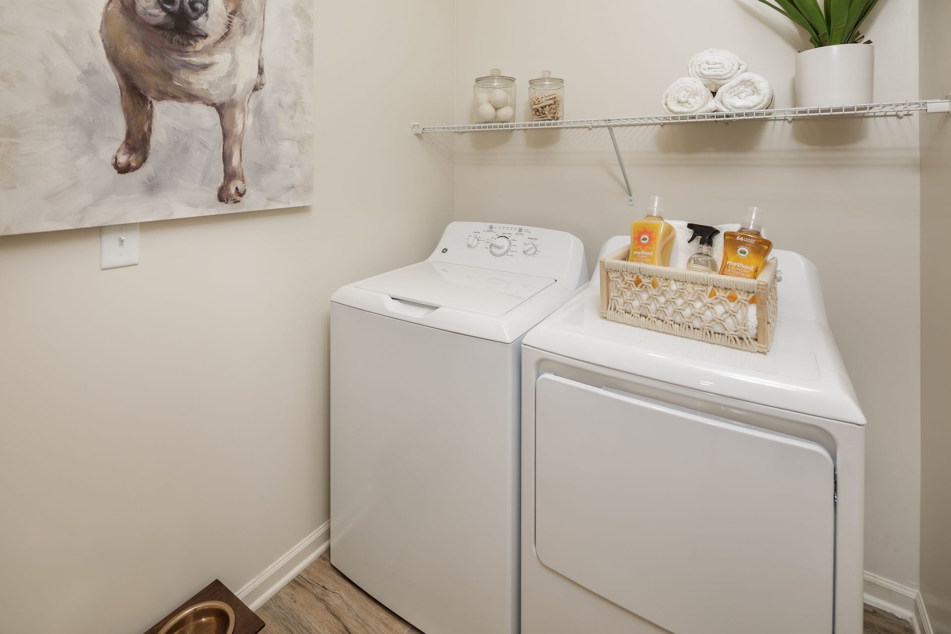 Laundry room with a white washer and dryer. Artwork, shelves with towels and jars.