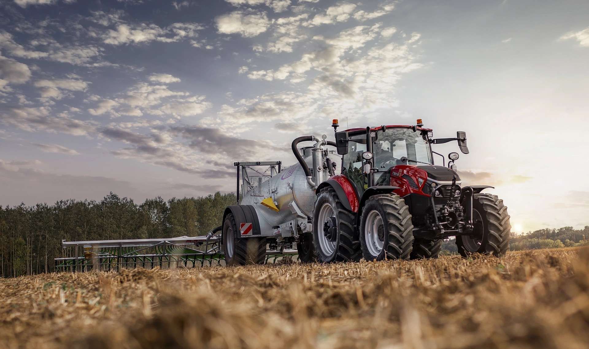 Red tractor pulling a large tank with field application equipment, in a harvested field under a cloudy sky.