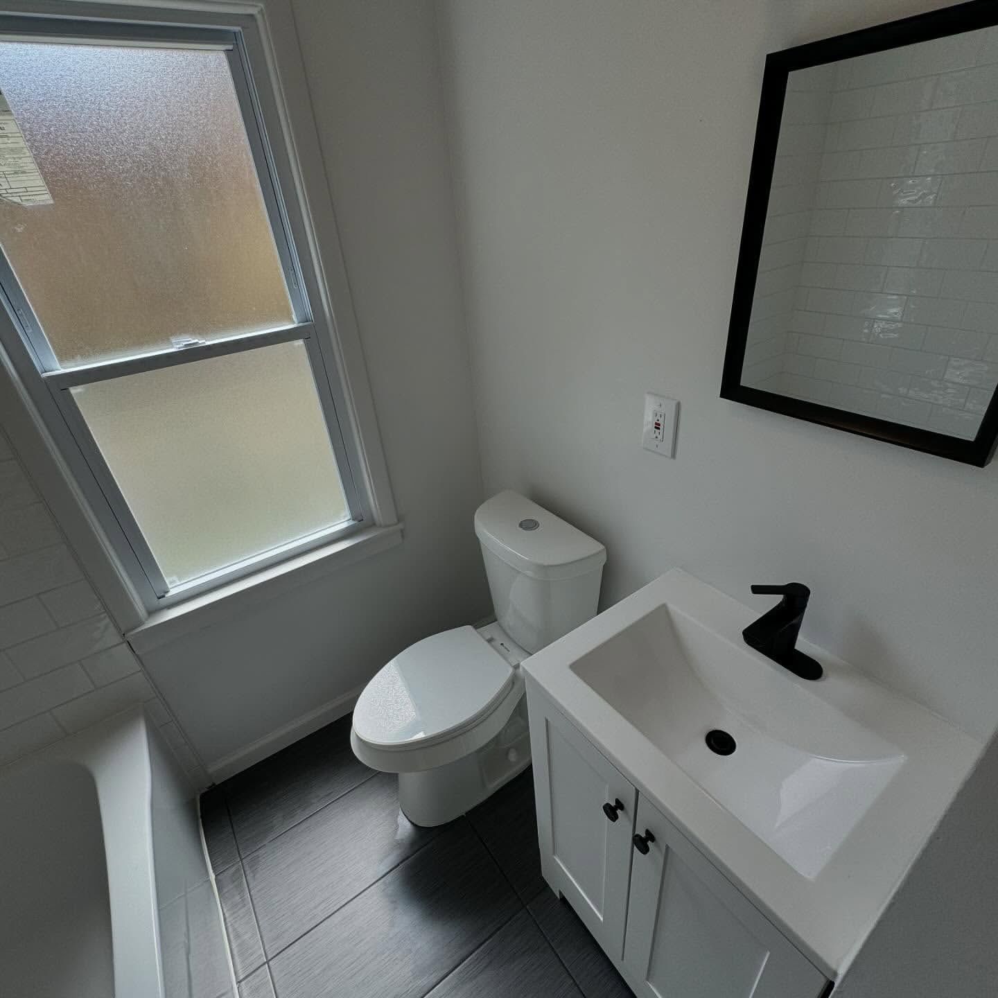 Small, modern bathroom with white vanity, black faucet, toilet, window, and dark gray floor tiles.