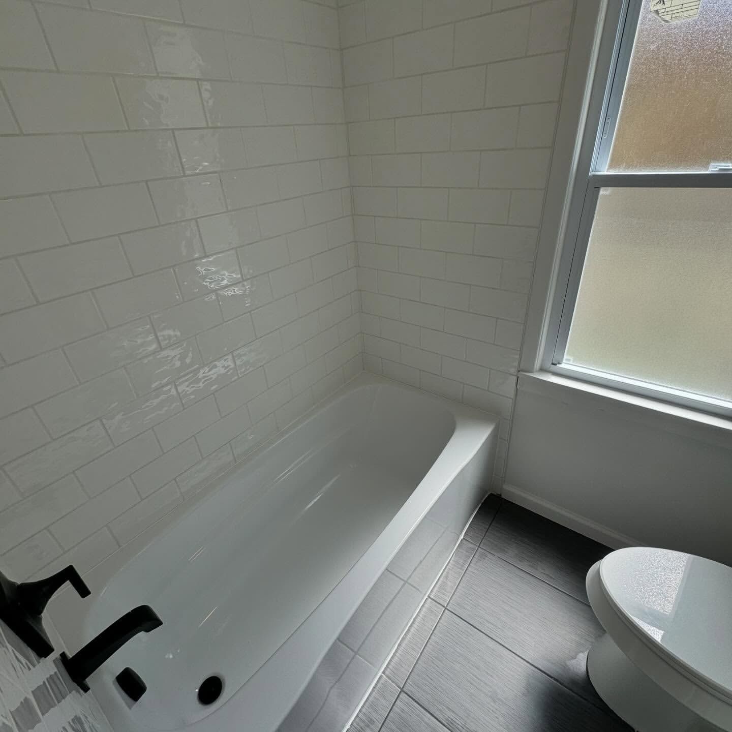 White-tiled shower/tub, dark floor, and toilet in a bathroom with a window.