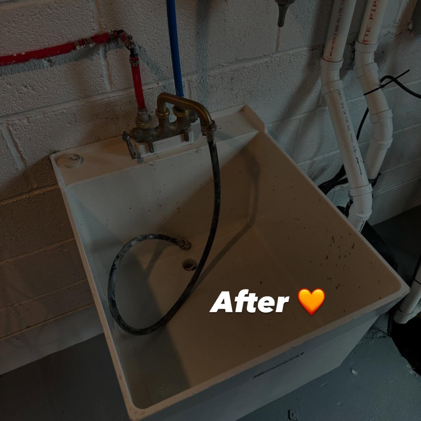 Laundry utility sink, white, against a gray cinder block wall, with water supply pipes and drain.