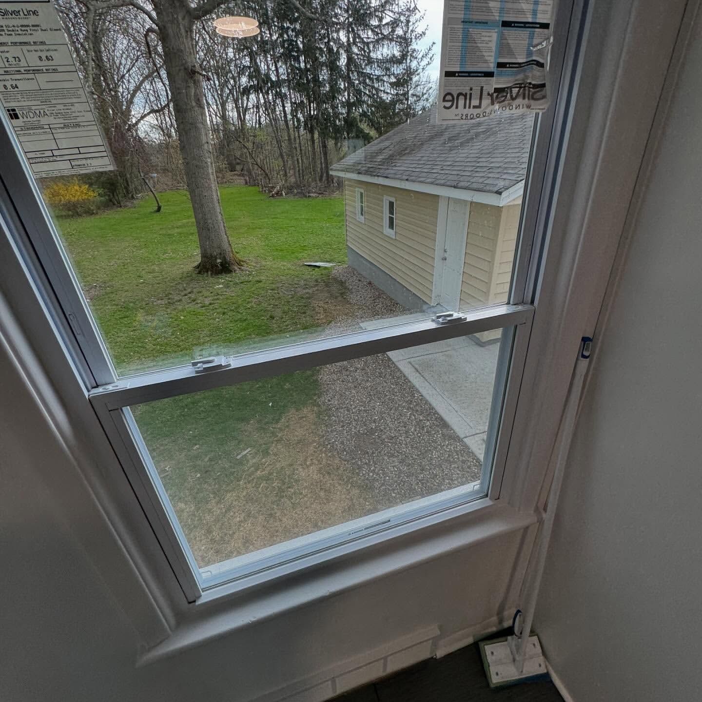 View through a white-framed window of a backyard with a small tan building, a tree, and green grass.