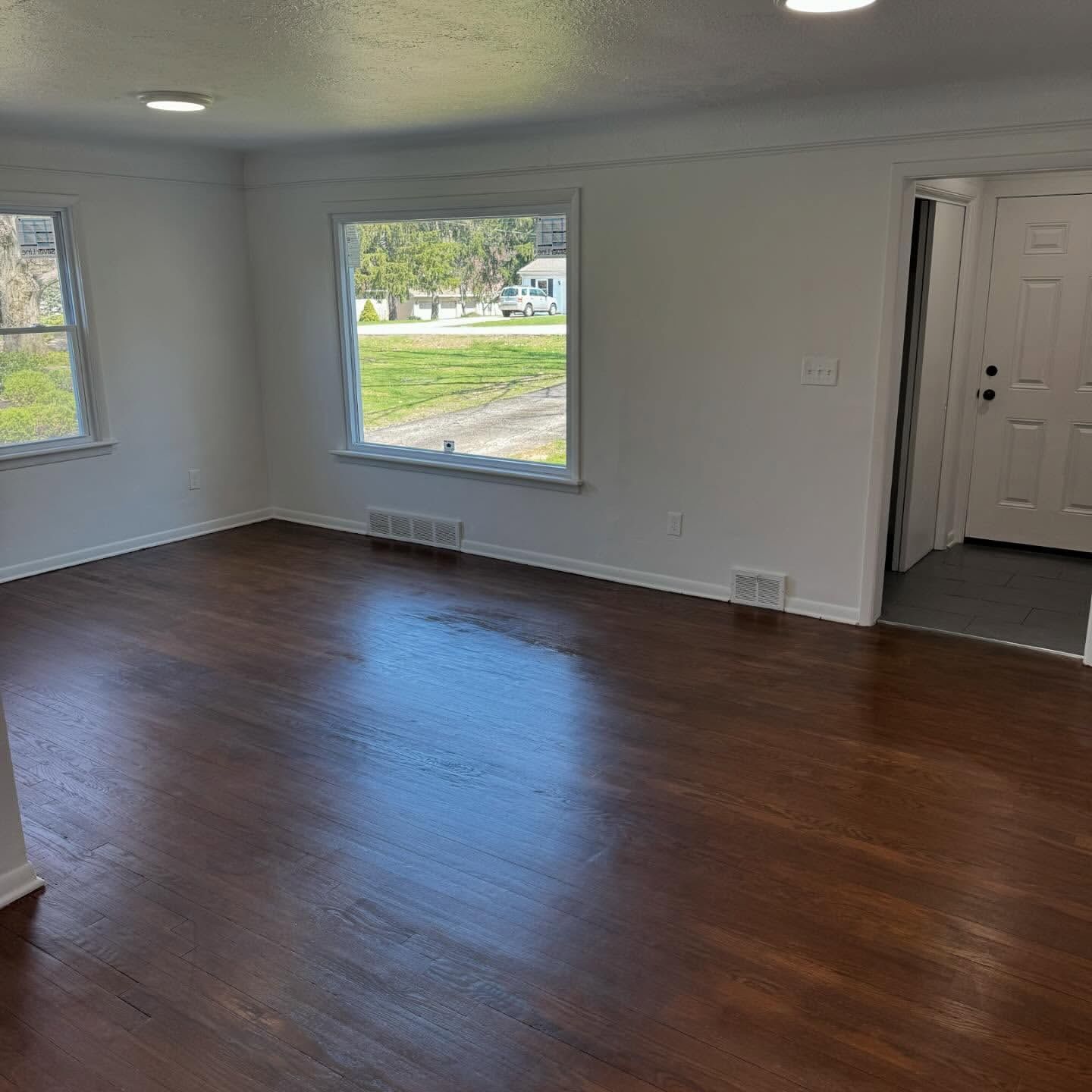 Empty living room with hardwood floors, white walls, and large windows overlooking a yard.
