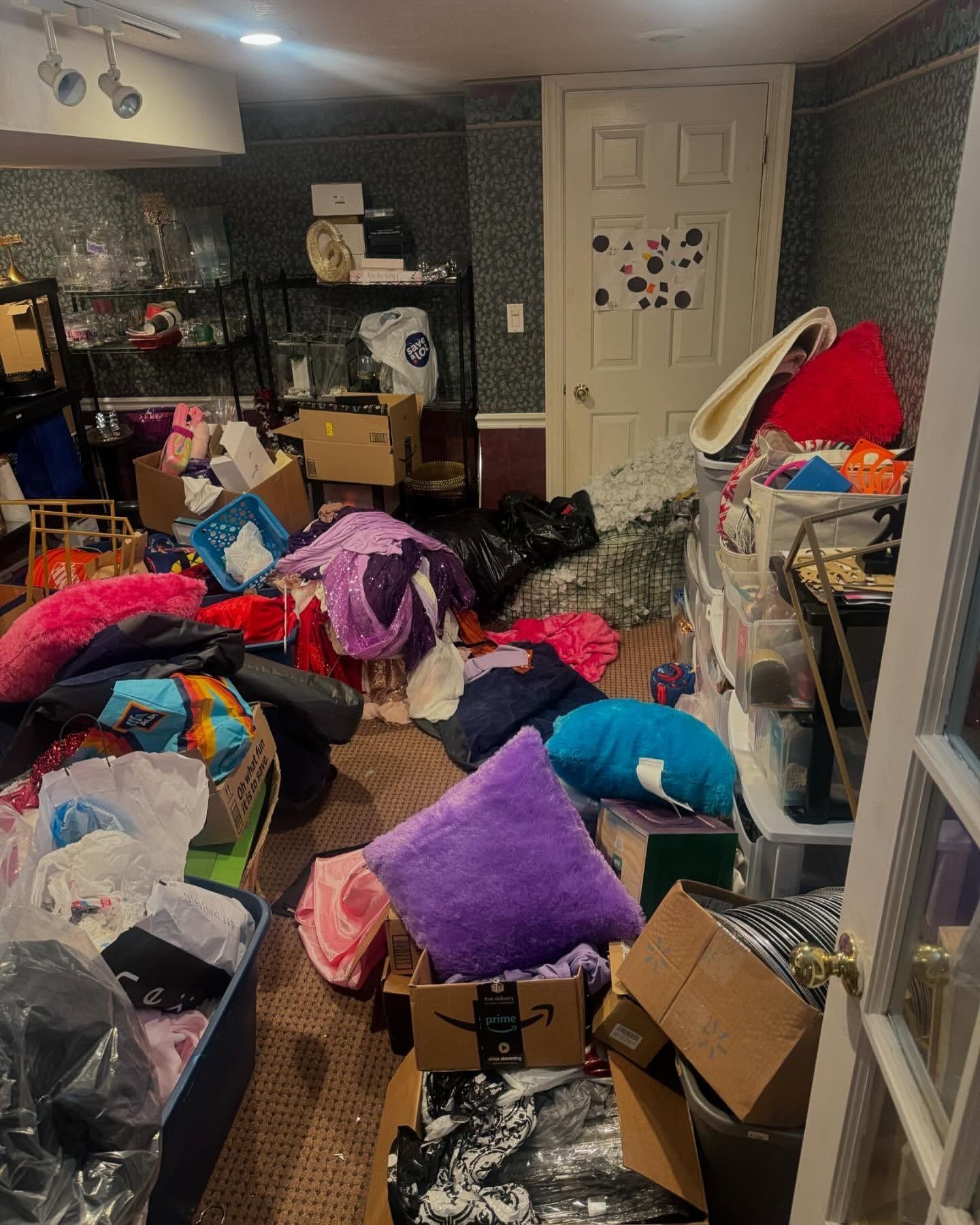 Cluttered basement with piles of clothes, boxes, and pillows near a door. Overwhelming disorganization.