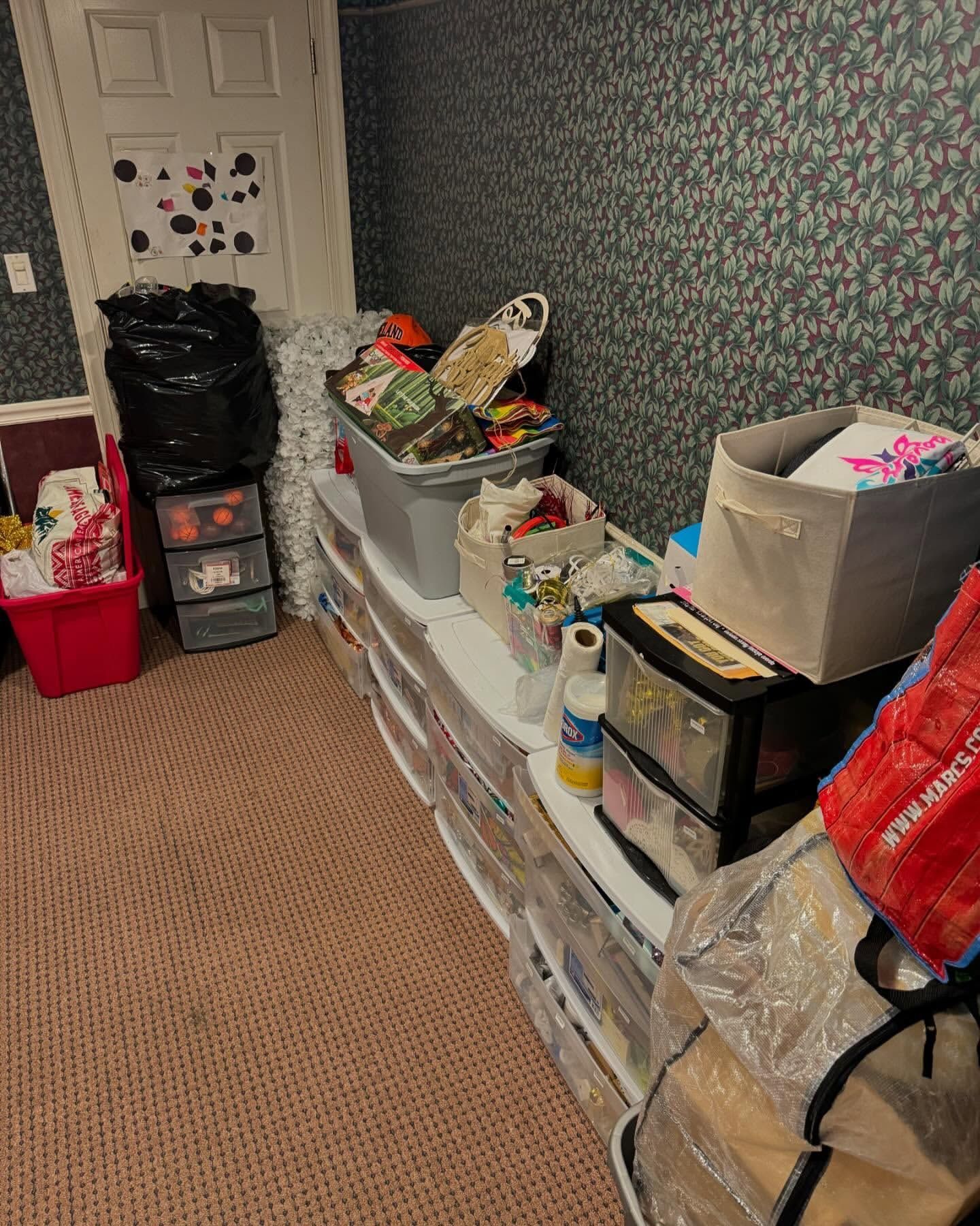 A cluttered storage area with shelves filled with bins and boxes. A trash bag and various items are present.