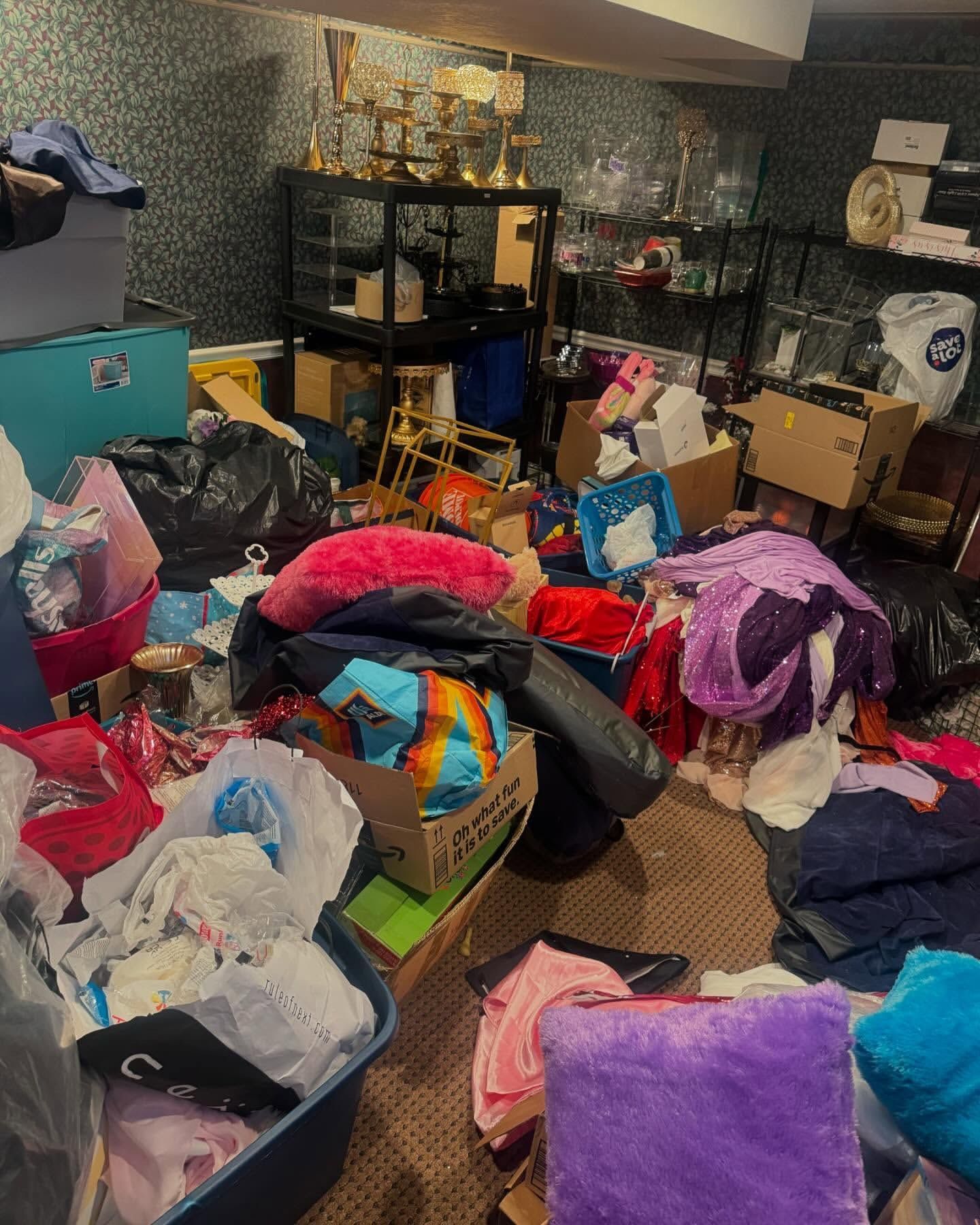 Cluttered basement: piles of clothes, boxes, and miscellaneous items on the floor near shelves displaying trophies and glassware.