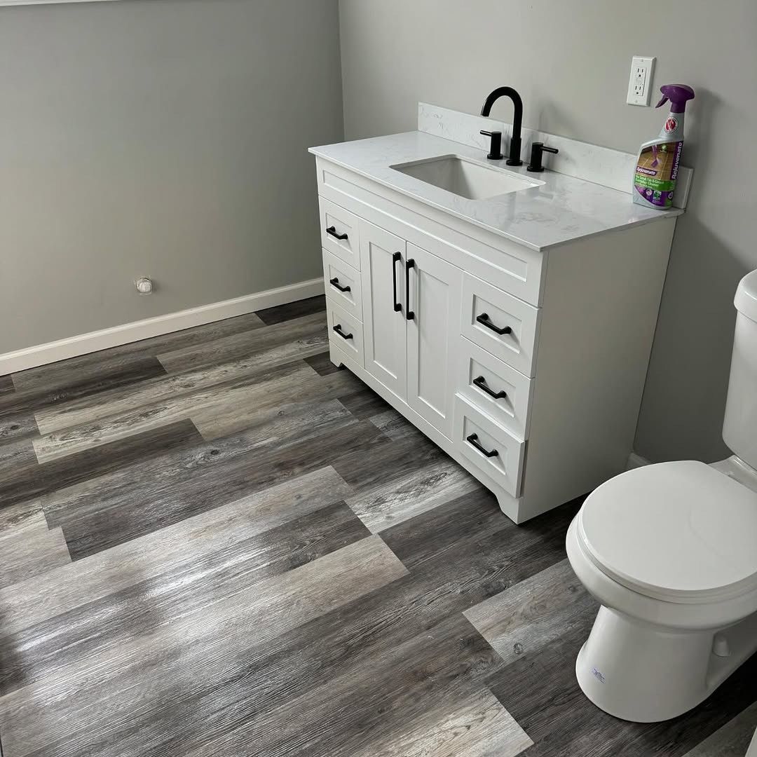 White bathroom vanity with black fixtures, next to a toilet. Gray wood-look flooring.