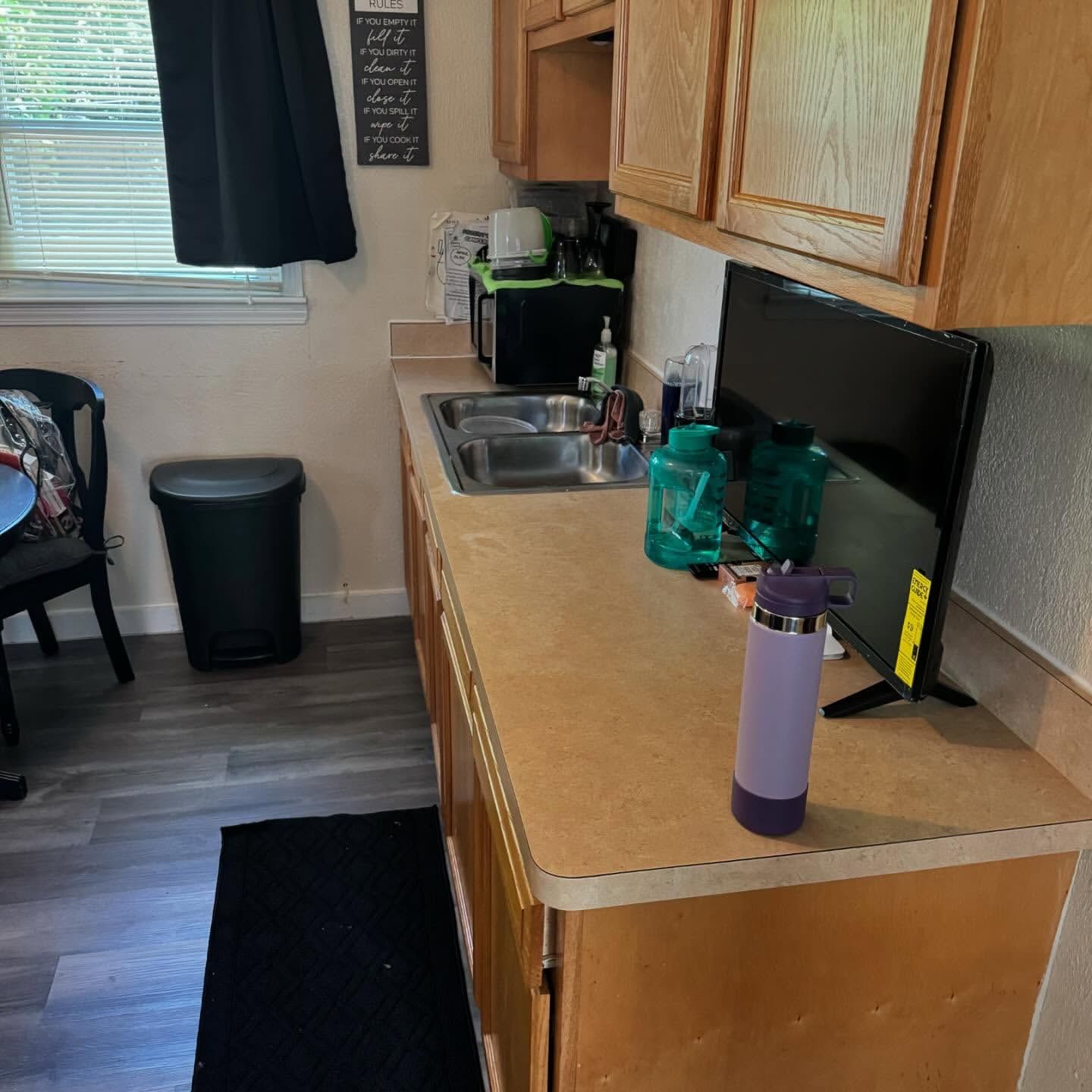 Small kitchen with a countertop, cabinets, sink, and a TV, with various items on the counter and a window.