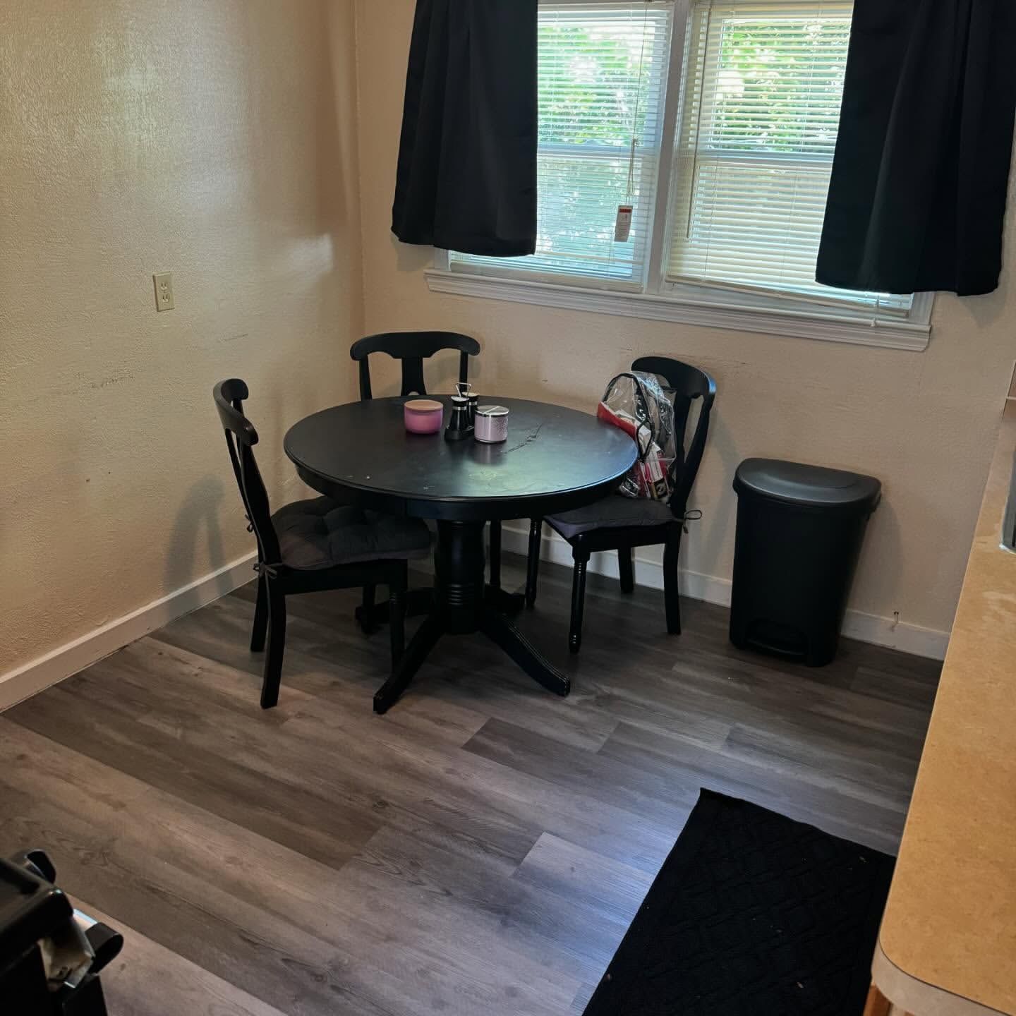 A small dining area with a round black table and three chairs, near a window with curtains.