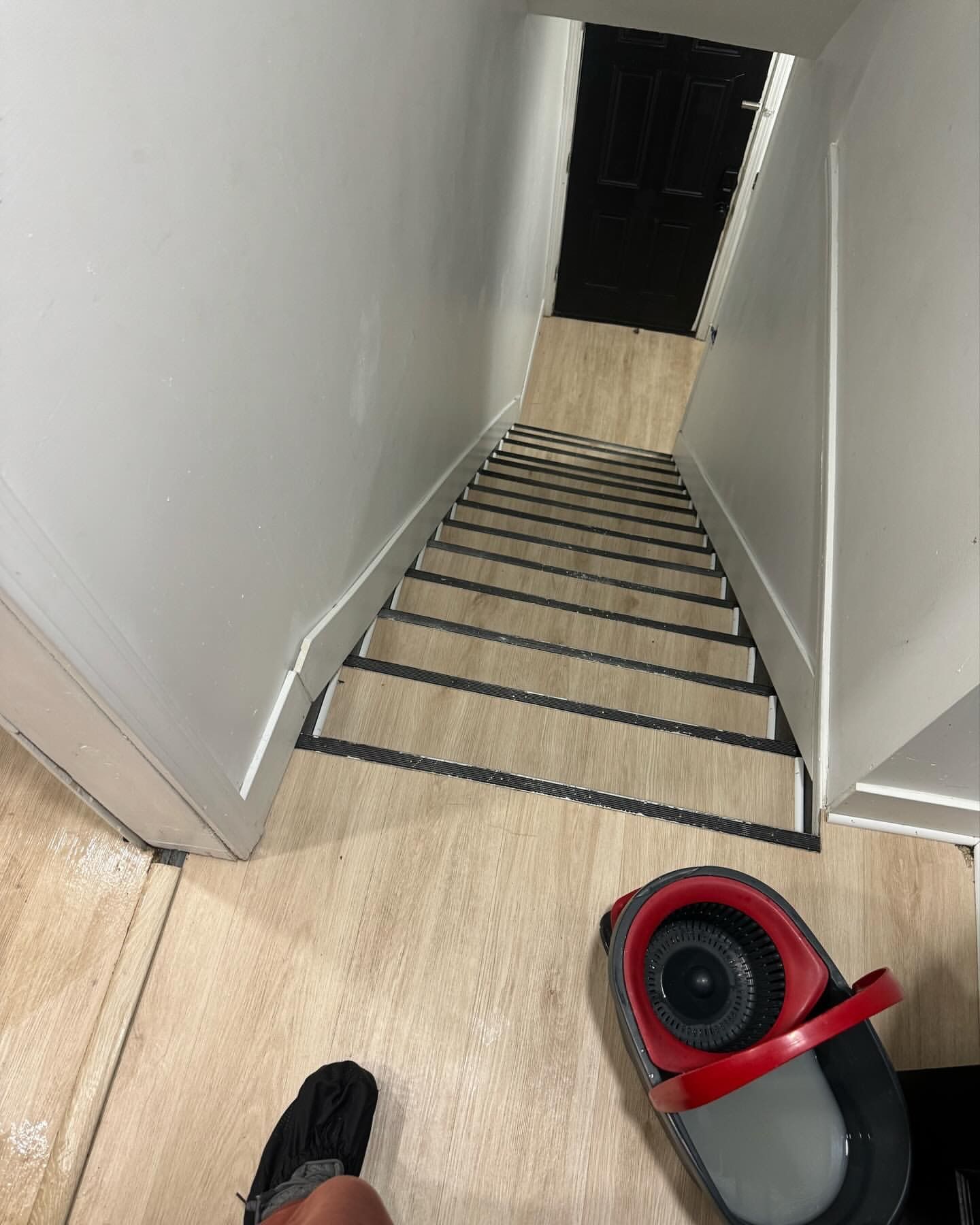 A person stands at the top of a staircase with a mop nearby. White walls and a black door are visible.
